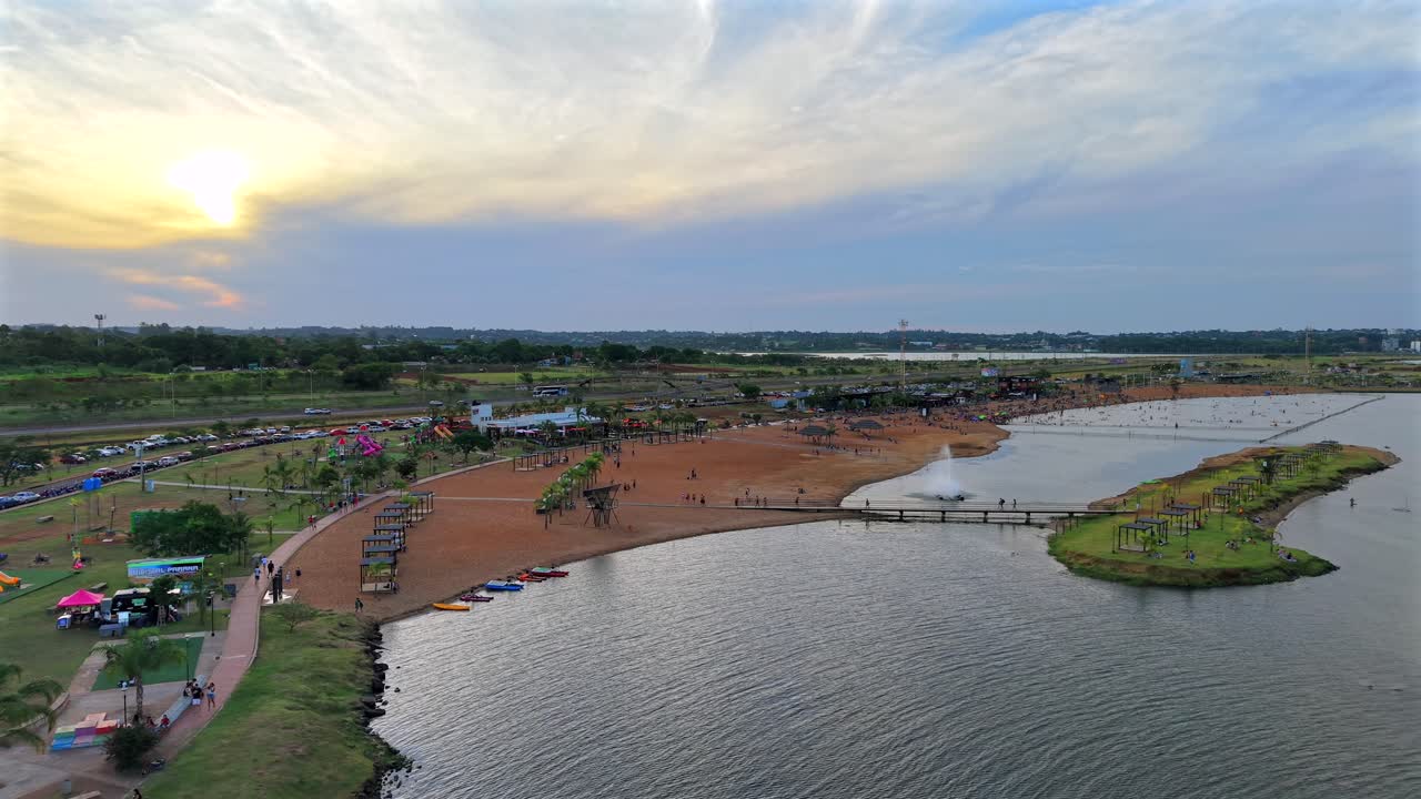 Balneario Costa Sur a Posadas, Misiones, Argentina, con litorale sabbioso, molo, kayak, paesaggio con palme e persone che si godono il fiume Paraná al tramonto, con il drone che si allontana.