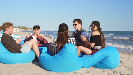 group of friends sitting on easychairs on the beach playing guitar and singing on a summer evening. slowmotion shot