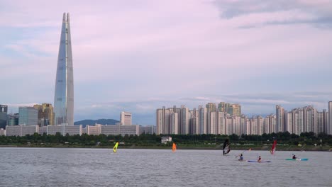 Seoul-Lotte-World-Tower-Landmark-building-from-the-bank-of-Han-river-at-Purple-colorful-sunset,-people-kayaking-and-riding-windsurfing-in-foreground