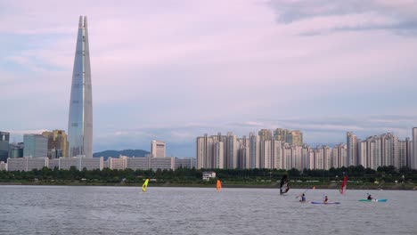 Edificio-Emblemático-De-La-Torre-Mundial-Lotte-De-Seúl-Desde-La-Orilla-Del-Río-Han-Al-Atardecer-De-Color-Púrpura,-Gente-Haciendo-Kayak-Y-Montando-Windsurf-En-Primer-Plano