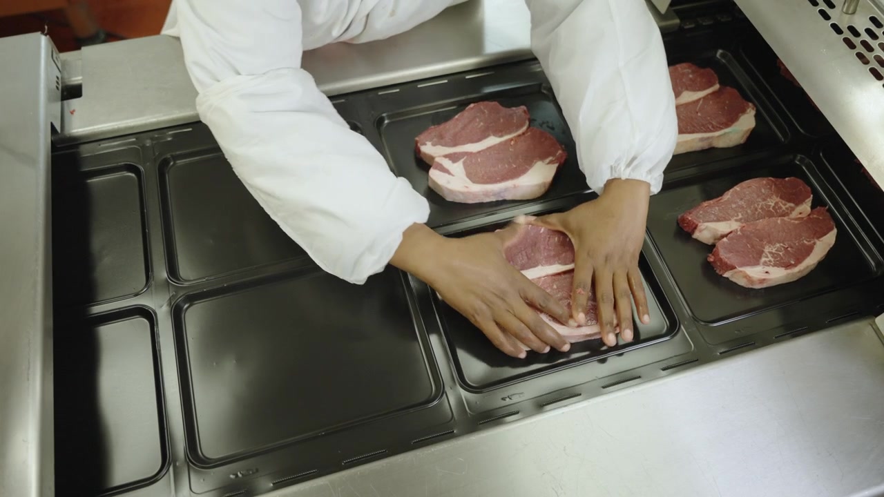 Gloved hands arrange raw beef steaks into black plastic trays on an automated packing line in a modern meat facility.
