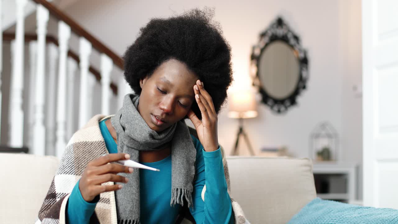 Close up of African American female wearing a scarf, sitting at home and holding thermometer