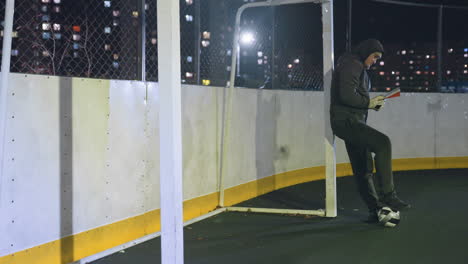 side view of man in hoodie leaning against metallic goalpost reading book outdoors at night with soccer ball underfoot, illuminated by urban city lights