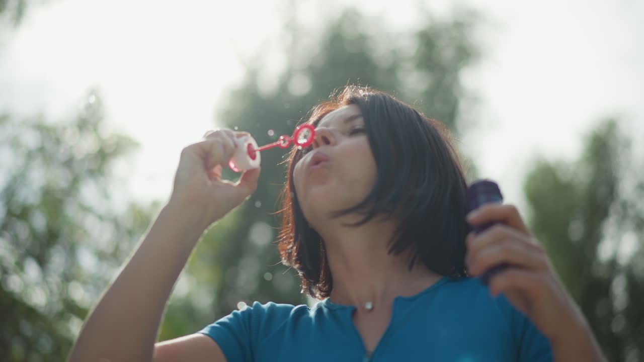 Blanke vrouw die buiten bellen blaast, zacht zonlicht door bomen, blauw shirt, langzame, bewuste ademhaling en zachte glimlach, sereen moment alleen met kleine, iriserende bellen die ronddrijven, ondiepe diepte