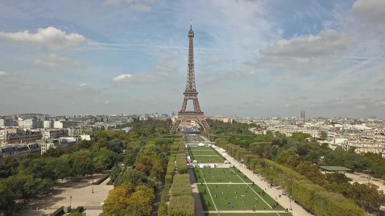 En droneoptagelse stiger op over Champ De Mars, der viser Eiffeltårnet på en solrig dag