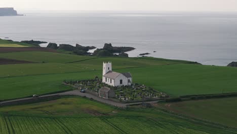 Toma-Aérea-En-Círculos-De-Una-Iglesia-Costera-Y-Campos-Verdes-Con-Ovejas-En-Irlanda-Del-Norte