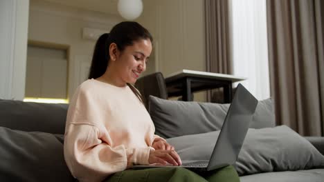 happy brunette girl in a pink jacket and green pants sits on a gray sofa and types on her laptop while working remotely at home happy during the day