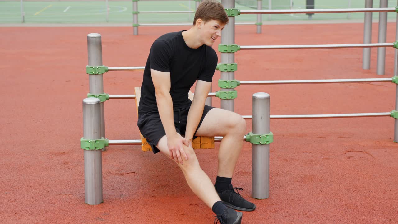Young man experiencing knee pain during outdoor workout, sitting on a bench at a calisthenics park resting and gently touching his injured leg while feeling discomfort, static camera slow motion shot