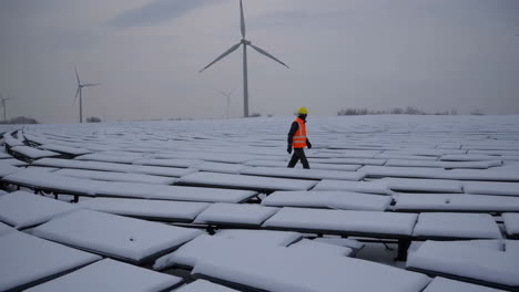 worker inspecting snow-covered solar panels with wind turbines in the background