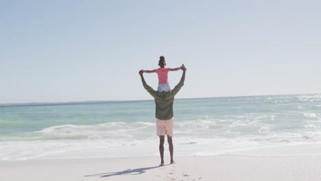 Afroamerikanischer-Vater-Trägt-Tochter-Mit-Ausgebreiteten-Armen-Am-Sonnigen-Strand