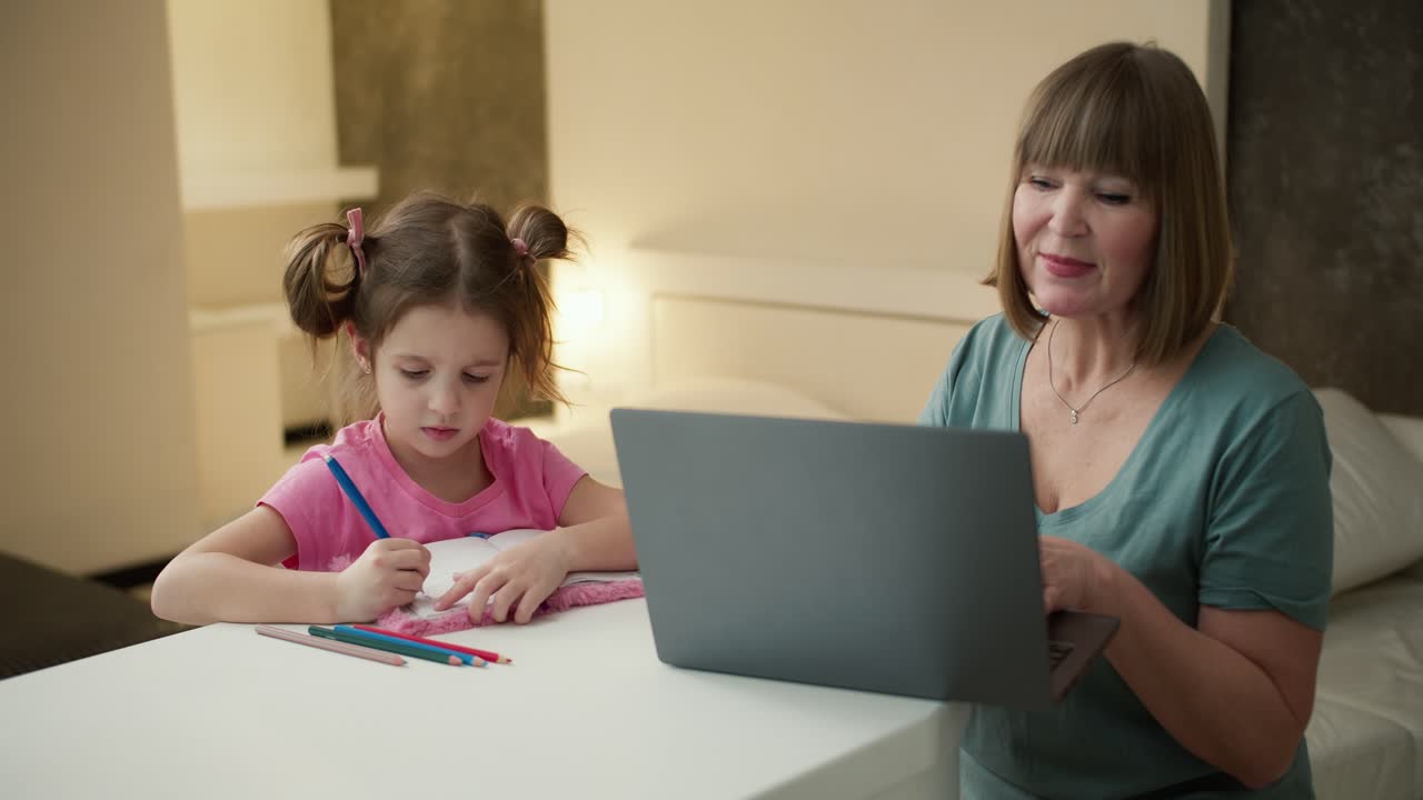 mujer anciana usando una computadora portátil mientras su nieta dibuja en el escritorio cercano