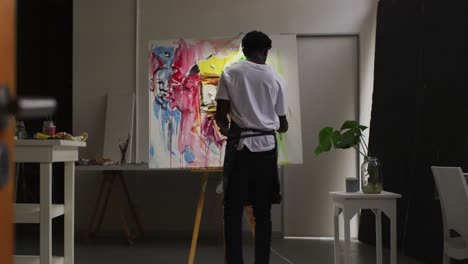 Rear-view-of-african-american-male-artist-wearing-apron-painting-on-canvas-at-art-studio