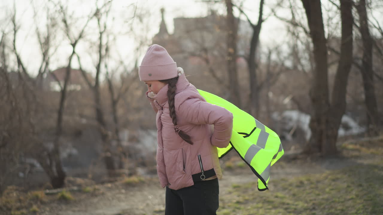 Woman in pink winter jacket and knit hat putting on bright yellow reflective safety vest outdoors in park area, preparing for volunteer or work activity on chilly day with trees and buildings in background