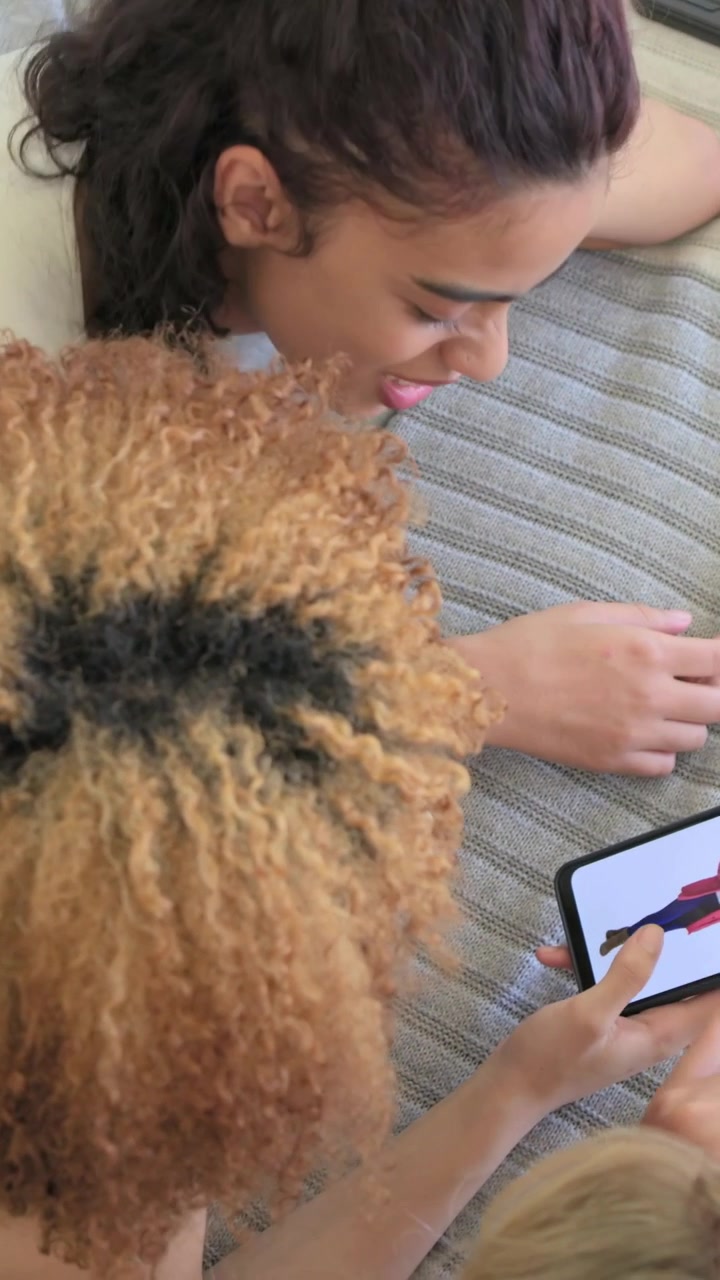 Vertical video: Smiling woman in sleeveless top lying on bed, friend tapping phone showing outfits