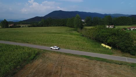 White-BMW-Car-Driving-Along-Green-Cornfields-In-Hostalets-De-Bas-In-Catalonia,-Spain