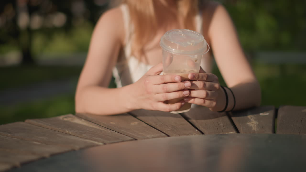 Giovane donna bianca che tiene in mano una bevanda in un parco soleggiato a un tavolo di legno, primo piano delle mani che stringono un bicchiere di plastica con una bevanda ghiacciata, calda luce dorata, profondità di campo ridotta, sfondo verde sfocato, rilassata