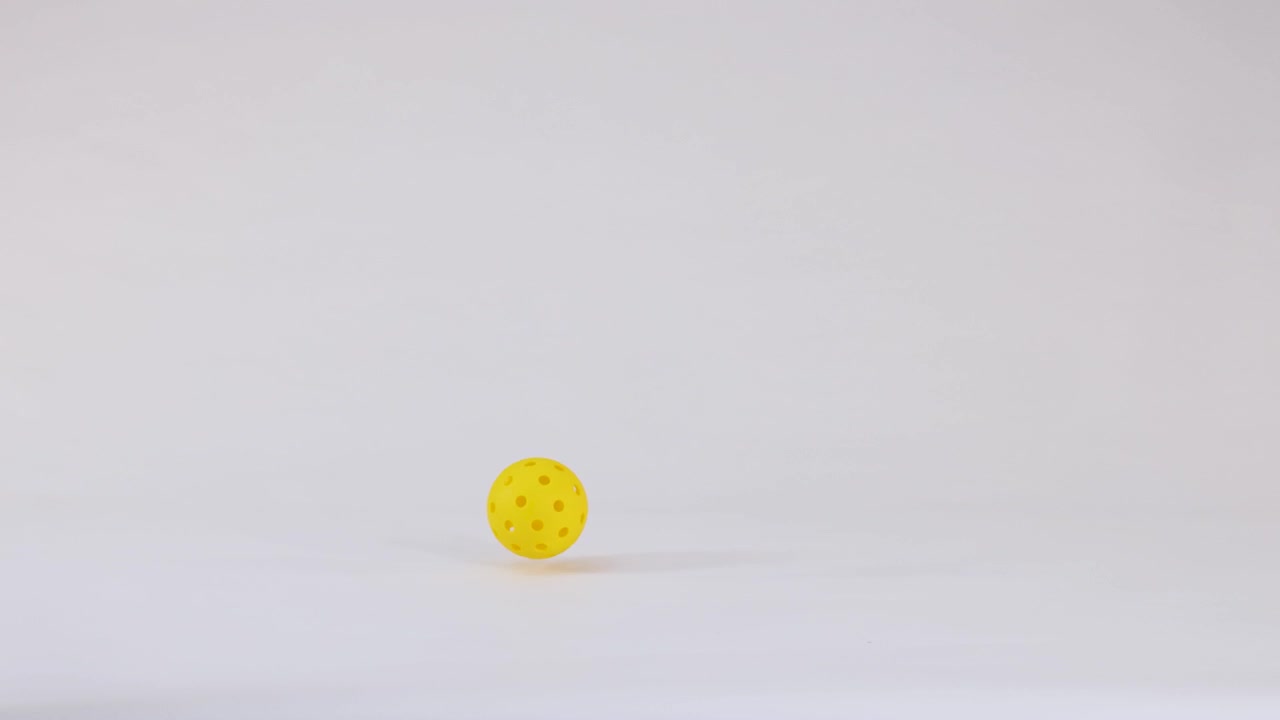 A single yellow pickleball falls into frame and bounces against a clean white studio background