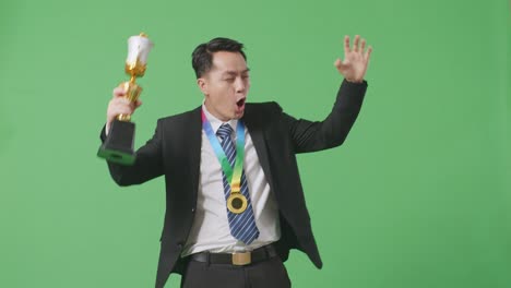 asian business man in a suit and tie with a gold medal looking at a gold trophy then celebrating winning as the first winner on green screen background in the studio