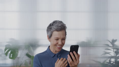 Retrato-Feliz-Mujer-De-Negocios-De-Mediana-Edad-Usando-Video-Chat-De-Teléfono-Inteligente-Saludando-Con-La-Mano-Beso-Disfrutando-De-La-Comunicación-Por-Teléfono-Móvil-En-La-Oficina-Cámara-Lenta