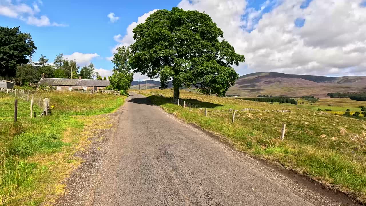 A vehicle travels along a narrow rural road in Clova, Angus, Scotland, passing stone walls, fields, and hills under bright daylight with dynamic clouds