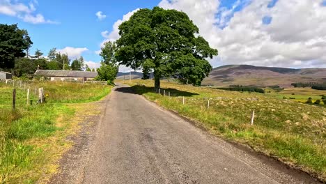 a vehicle travels along a narrow rural road in clova, angus, scotland, passing stone walls, fields, and hills under bright daylight with dynamic clouds