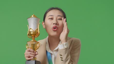 close up of asian business woman in a suit with a gold medal holding a gold trophy and yelling positively with hand over mouth on green screen background in the studio