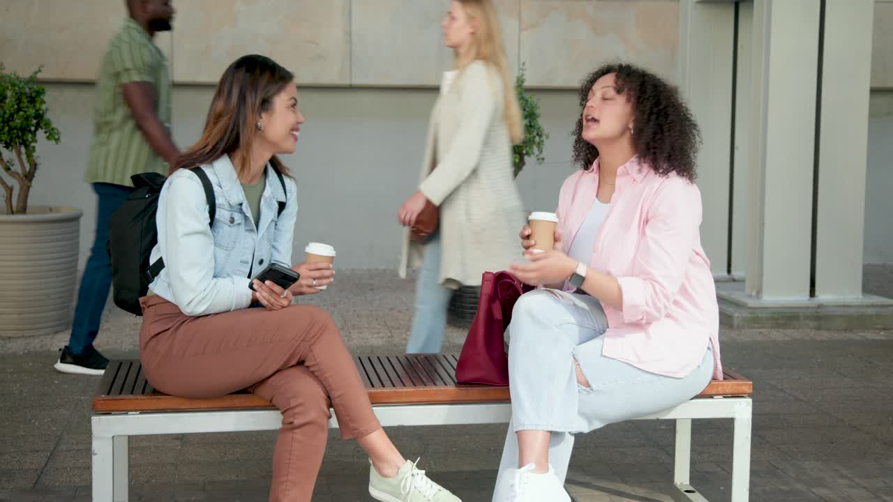 Two adult women left woman showing phone on bench outside while right sipping coffee sharing laugh