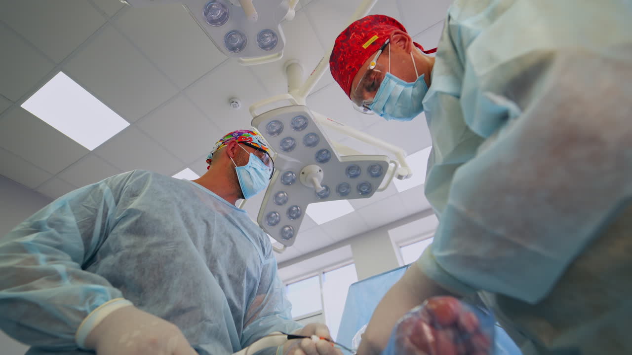 Male doctors in protective uniform using equipment operating the patient. Cooperative surgeons talk, advise and discuss the process. Low angle perspective from patient's view.