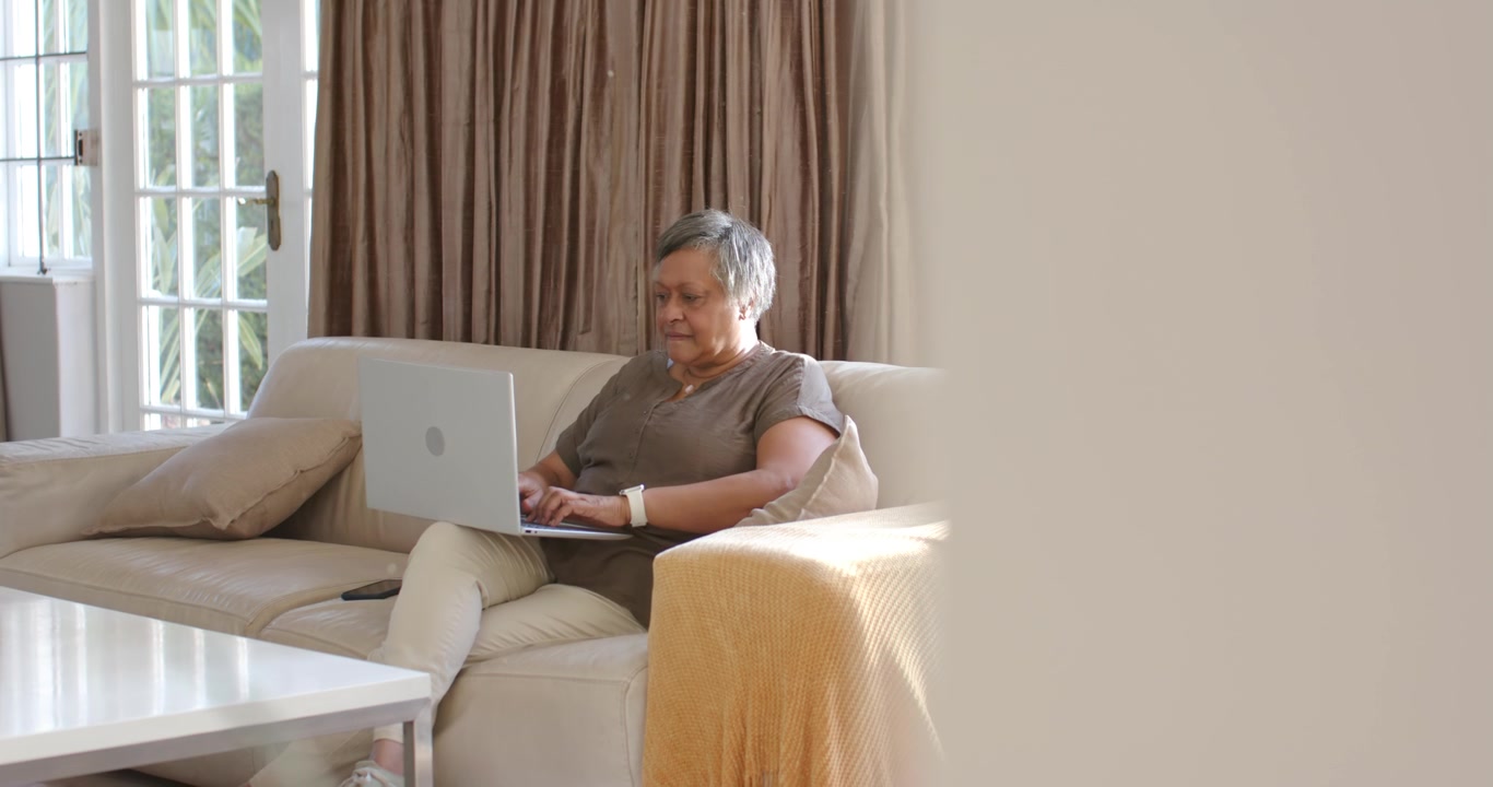 Senior woman of African descent sitting on sofa with laptop open on lap, typing and working