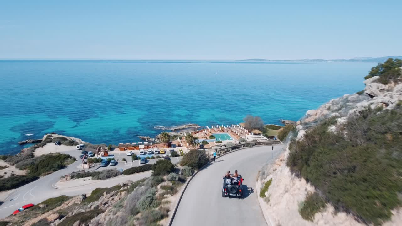 Stunning aerial footage of jeep driving towards a beach with dancing girls having fun in the SUV and blue turquoise water in the background - 4k drone shot following the car