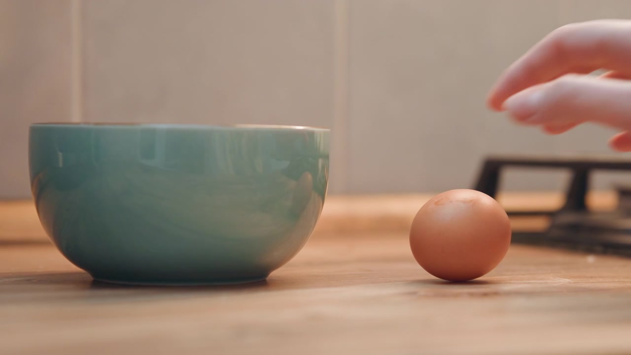 Hand placing egg into ceramic bowl, warm wooden counter, gas stove backdrop, gentle motion of hand setting brown egg beside bowl, closeup domestic scene, breakfast preparation, tactile texture