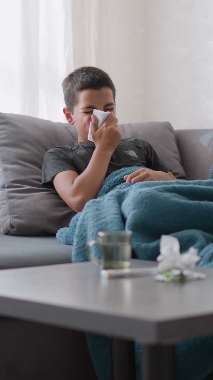 Sick boy sitting on couch covered with blue blanket, sneezing into tissue, looking at it with used tissue and glass cup on table beside him, cozy indoor setting with natural light