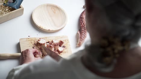 caucasian senior woman with braid, cutting octopus tentacles on cutting board with a knife, preparing them for cooking, shot from the back