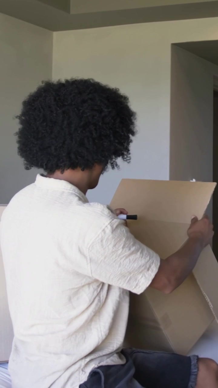 Vertical video: Lifting flat cardboard, man seated in cream shirt assembling box on bed with marker