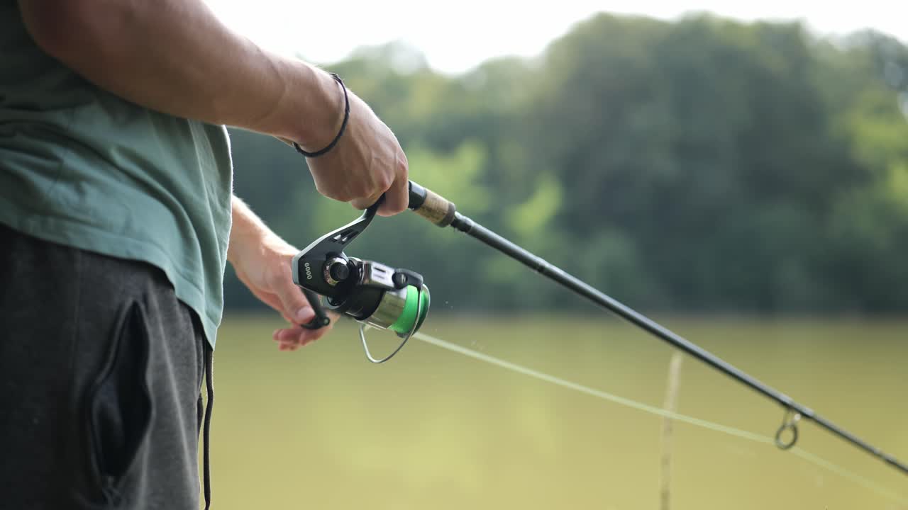 Male angler stands on the shore of very cloudy lake and turns the fishing rod winch to reel in the line