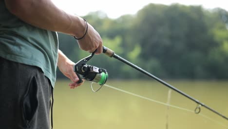 Male-angler-stands-on-the-shore-of-very-cloudy-lake-and-turns-the-fishing-rod-winch-to-reel-in-the-line