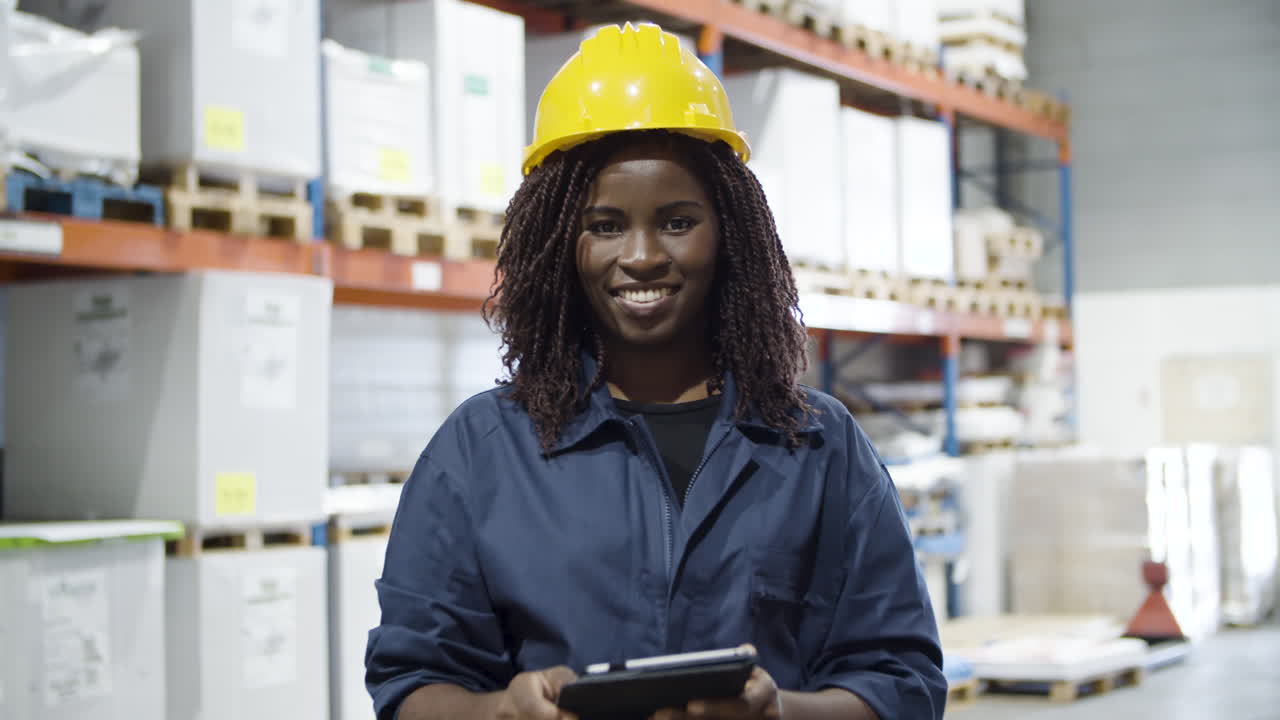 Happy African American female employee of logistic company standing in warehouse and smiling at the camera