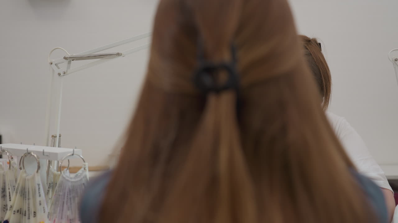 Nail technician wearing mask and gloves carefully working on client nails using scissors and blue brush under white lamp, client with blonde hair tied back with black clip seated at manicure station