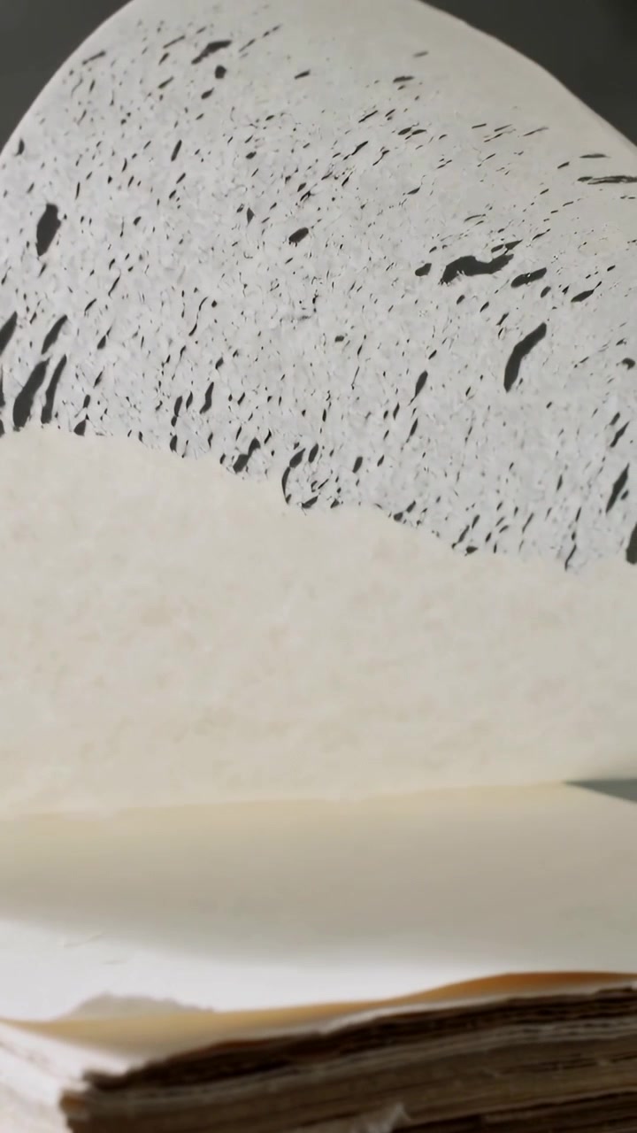 Video verticale: Foglio superiore trasparente maculato che si stacca, rivelando bordi marroni invecchiati su una scrivania, parete grigia.