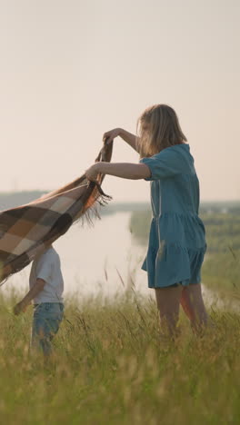 a woman in a blue dress holds a large plaid scarf high as it catches the wind on a lush grassy hilltop beside a lake. two children in white shirts play nearby,enjoying the gentle breeze and open space