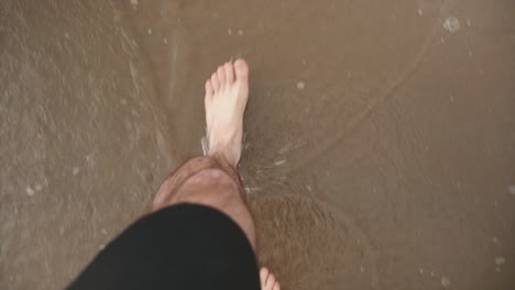 a caucasian mans feet walking on a beach as the tide periodically comes in