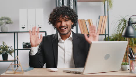 businessman working on laptop smiling friendly at camera and waving hands gesturing hello at office