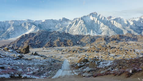 Majestic gravel road leading to snowy mountain range during snowfall, aerial fly forward view