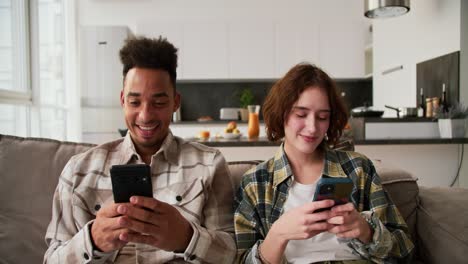 A-happy-man-with-Black-skin-a-brunette-with-stubble-and-his-girlfriend-in-checkered-shirts-are-sitting-on-a-gray-sofa-and-looking-at-programs-in-their-smartphones-and-Social-Media-sitting-on-the-sofa-in-a-modern-apartment