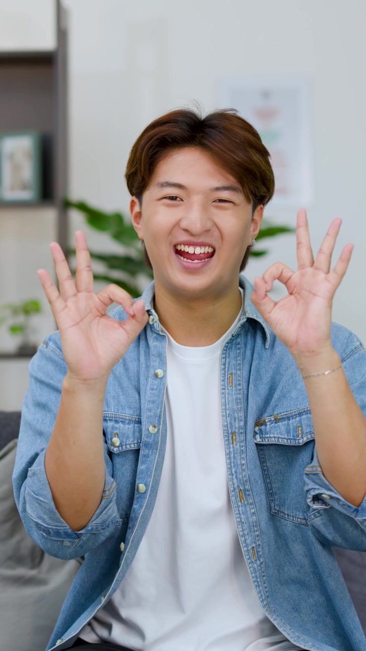 Un giovane uomo asiatico allegro con una camicia di jeans fa doppi segni di OK mentre sorride.