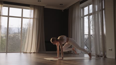 A-young-beautiful-woman-in-white-sportswear-stretches-in-a-gym-with-large-windows-in-slow-motion-in-the-sun's-rays