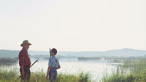 Rear-view-of-senior-man-and-his-grandson-standing-on-the-river-bank-and-talking-with-rods-in-hands