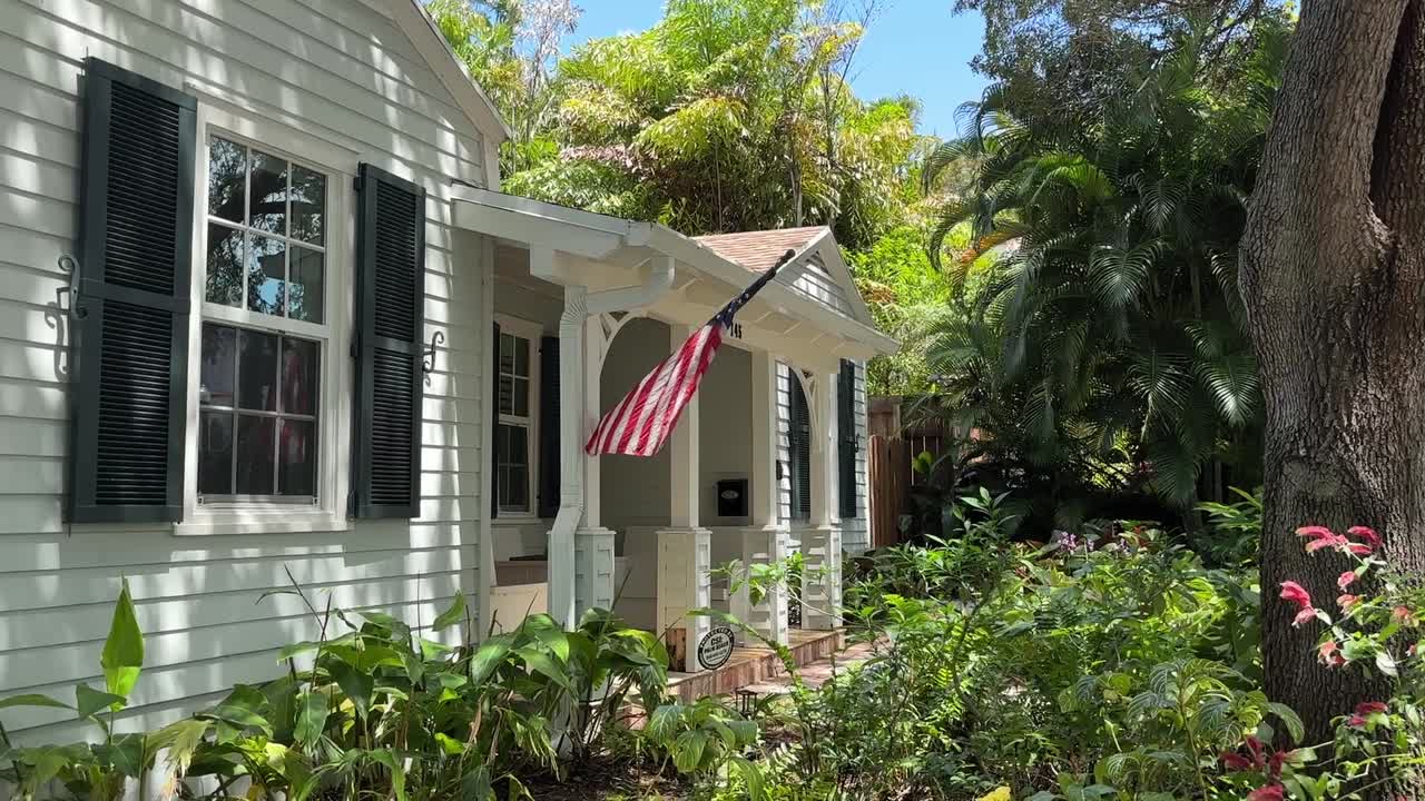 A white wooden house with a front porch and garden surrounded by tropical plants and trees is seen on a sunny day in Palm Beach, Florida