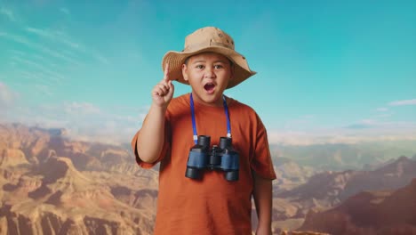 asian boy with a hat and binoculars thinking and looking around then raising his index finger while traveling at the top of mountain. boy researcher, travel tourism adventure concept