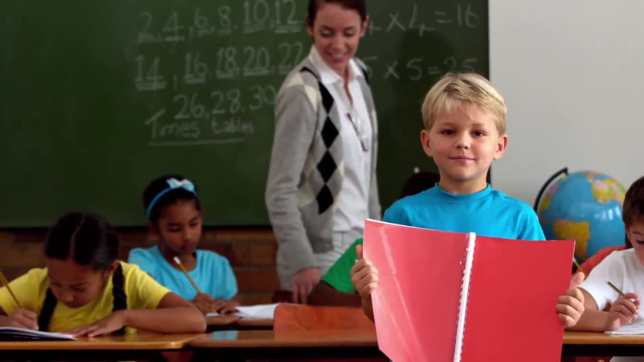 niño pequeño sosteniendo un bloc de notas rojo sonriendo a la cámara en el aula